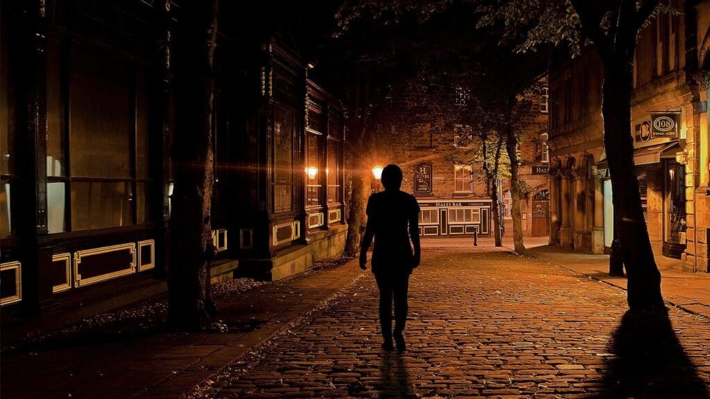 Silhouette of a person walking alone down a lamp-lit street at night